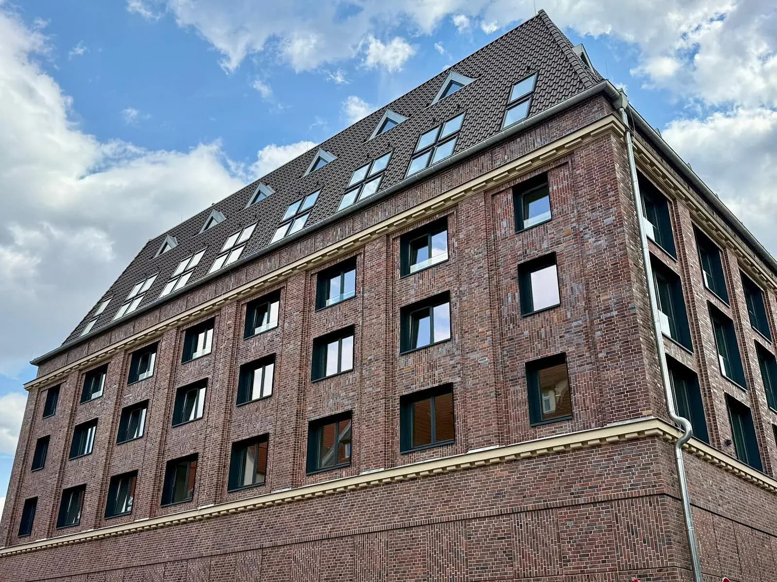Backsteinbau des MalzQuartiers von schräg unten: rotbraune Klinkerfassade mit hellen Gesimsen, dunklen Fensterrahmen und zwei Reihen Gauben im steilen Ziegeldach